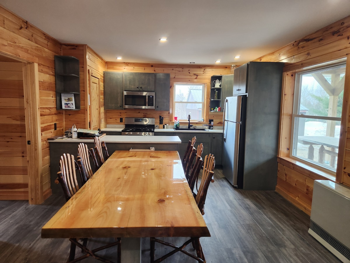 A spacious kitchen is highlighted by a large wooden dining table surrounded by six rustic chairs. Cabinets are crafted from dark wood, complementing stainless steel appliances. Natural light streams through the window, illuminating the room's wooden walls and modern fixtures.
