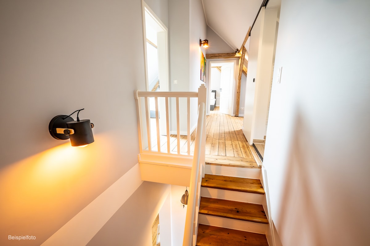 A well-lit hallway features wooden flooring and a staircase leading to the lower level. Soft wall sconces provide gentle illumination, while light-colored walls enhance the spacious feel. Doorways to adjacent rooms are visible along the corridor.