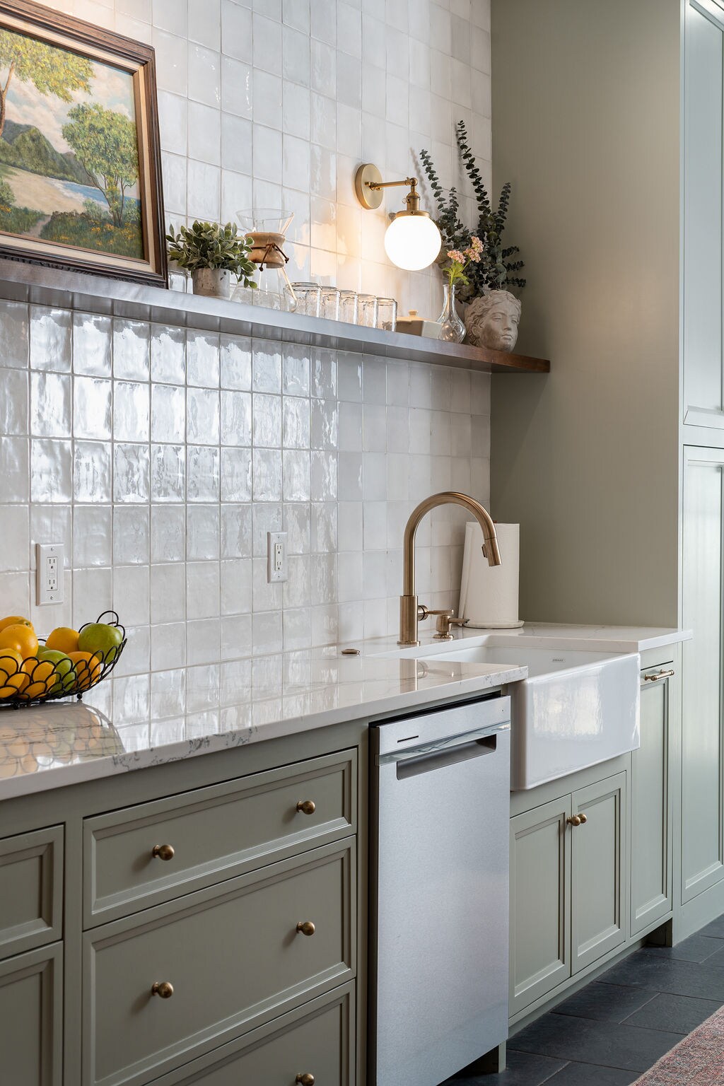 A modern kitchen showcases light green cabinetry with gold knobs, complemented by a white farmhouse sink. A marble countertop runs under a decorative shelf holding plants and glassware. Soft natural light illuminates the space, highlighting the textured wall tiles and a bowl with fresh fruit.