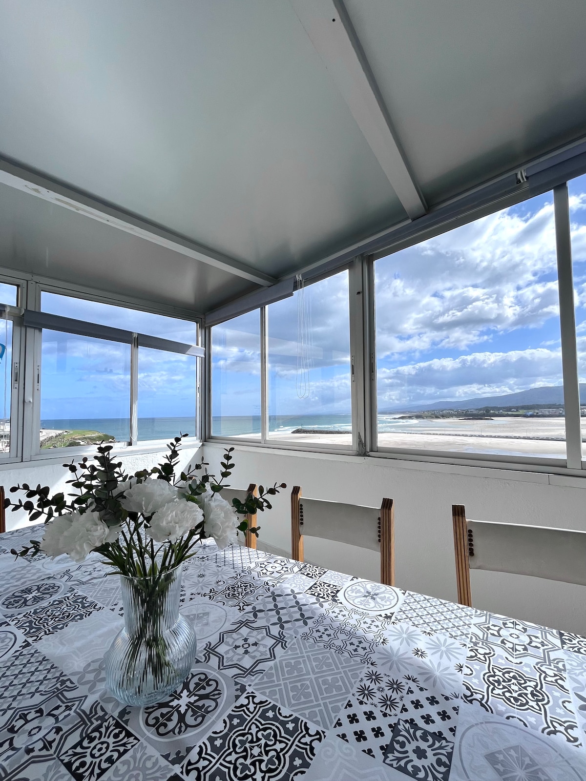A dining area is showcased with a patterned tablecloth and a vase of white flowers at the center. Large windows surround the space, providing views of the beach and ocean beyond, allowing natural light to fill the room.