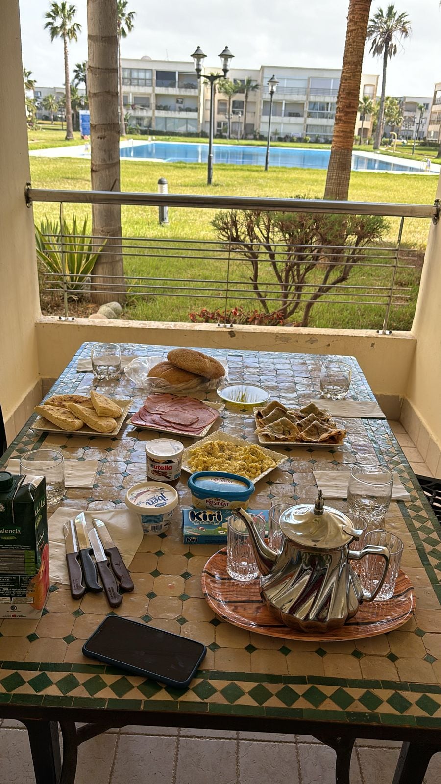 A table is set on a balcony overlooking a grassy area and a pool. The spread includes glassware, a teapot, a container of cream cheese, bread, and a selection of spreads. Palm trees and the distant pool are visible in the background.