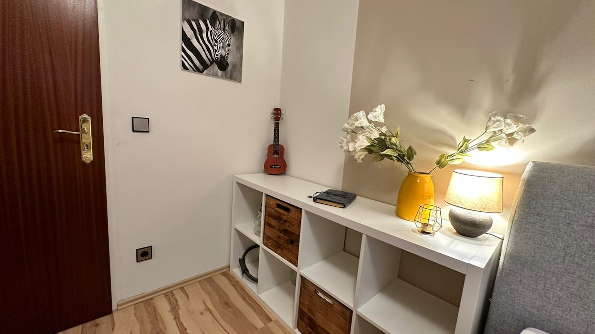 A corner of the room includes a white shelving unit with wooden drawers, complemented by a modern lamp and a decorative vase filled with flowers. A small guitar hangs on the wall beside a framed black-and-white zebra photograph, adding character to the space.