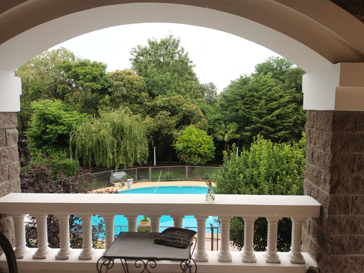 A balcony offers a view of a landscaped garden featuring lush greenery and tall trees. A swimming pool is visible in the foreground, surrounded by a fence. A small table with a cloth rests on the balcony, providing a cozy spot to enjoy the scenery.