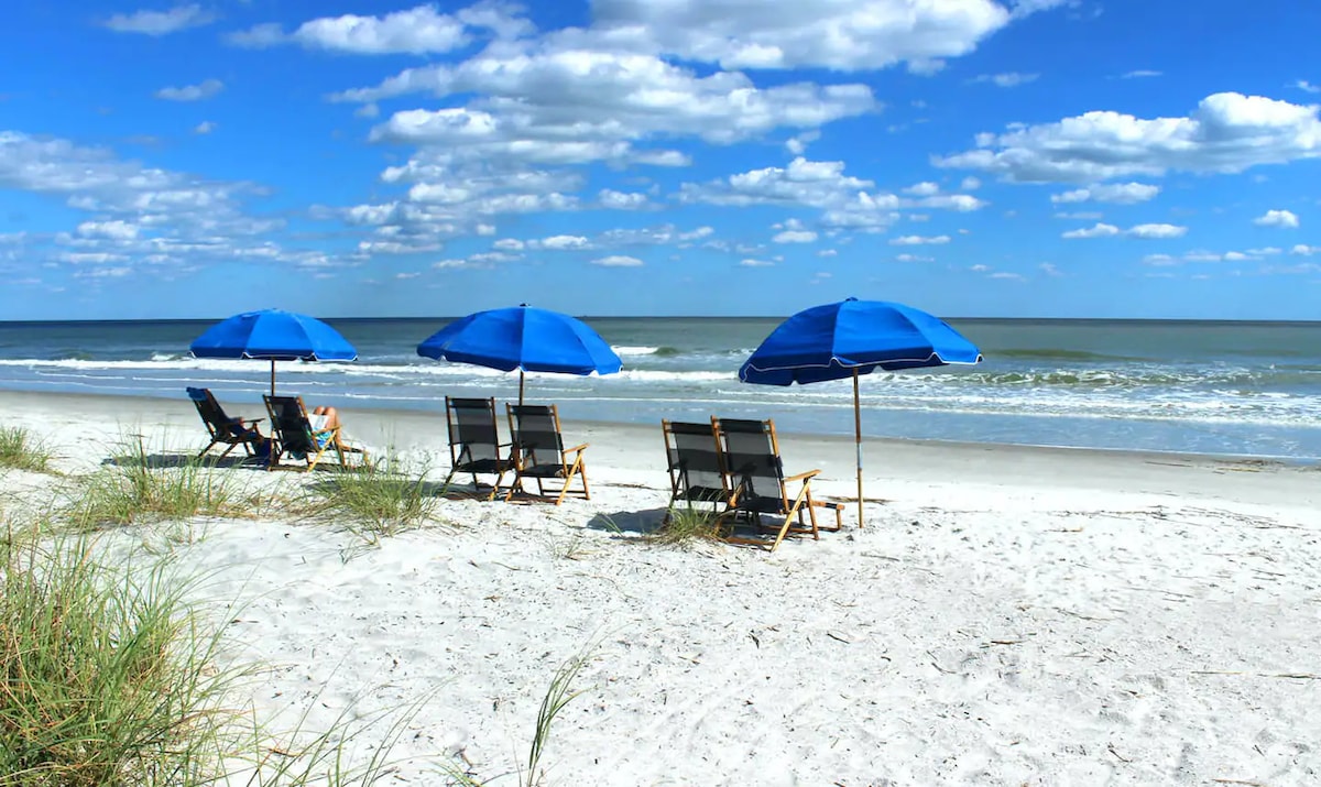 Four beach chairs are arranged under blue umbrellas on a sandy shore, with gentle waves lapping at the beach. A clear blue sky with scattered clouds stretches overhead, creating a serene coastal atmosphere.
