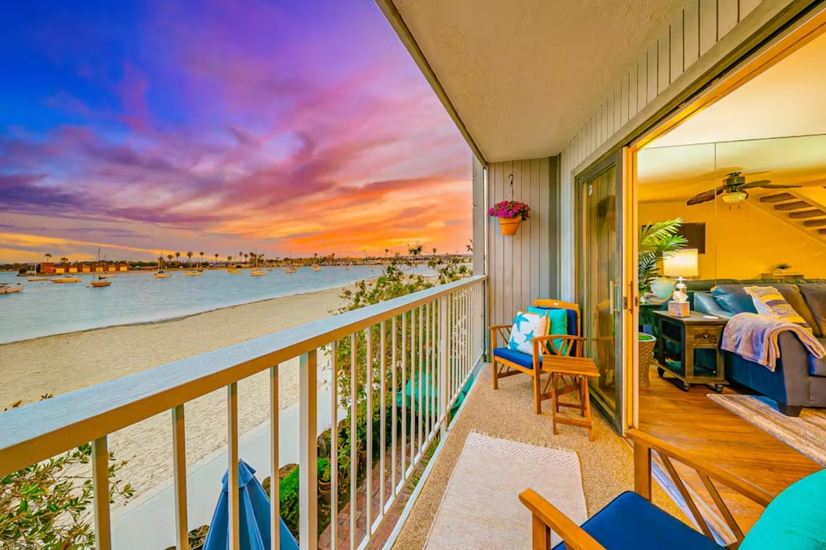 A private balcony offers scenic views of the bay during sunset, showcasing vibrant colors in the sky. Two comfortable chairs and a small table invite relaxation. A sliding door connects to the interior living space, enhancing the coastal living experience.