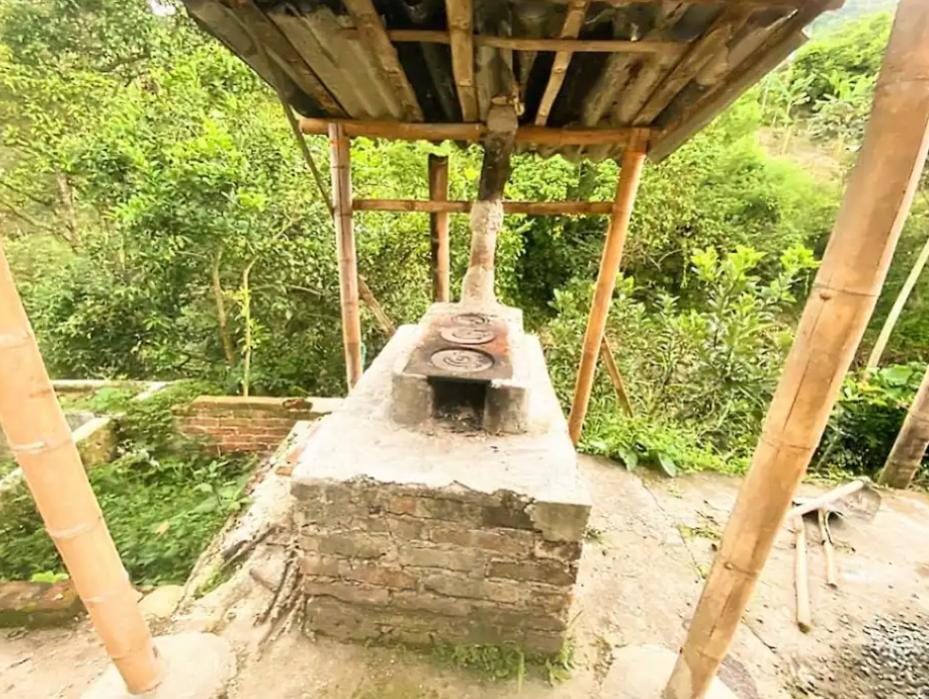 An outdoor cooking area is featured, consisting of a rustic stone and brick barbecue grill under a bamboo structure. The cooking surface includes two burners, surrounded by a natural green landscape filled with trees and vegetation.