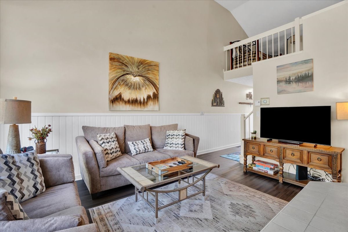 A spacious living area features a comfortable, light gray sofa adorned with patterned cushions. A wooden coffee table sits atop a soft area rug, complementing the warm-toned wall art above. Large windows allow natural light to fill the room, enhancing the inviting atmosphere.