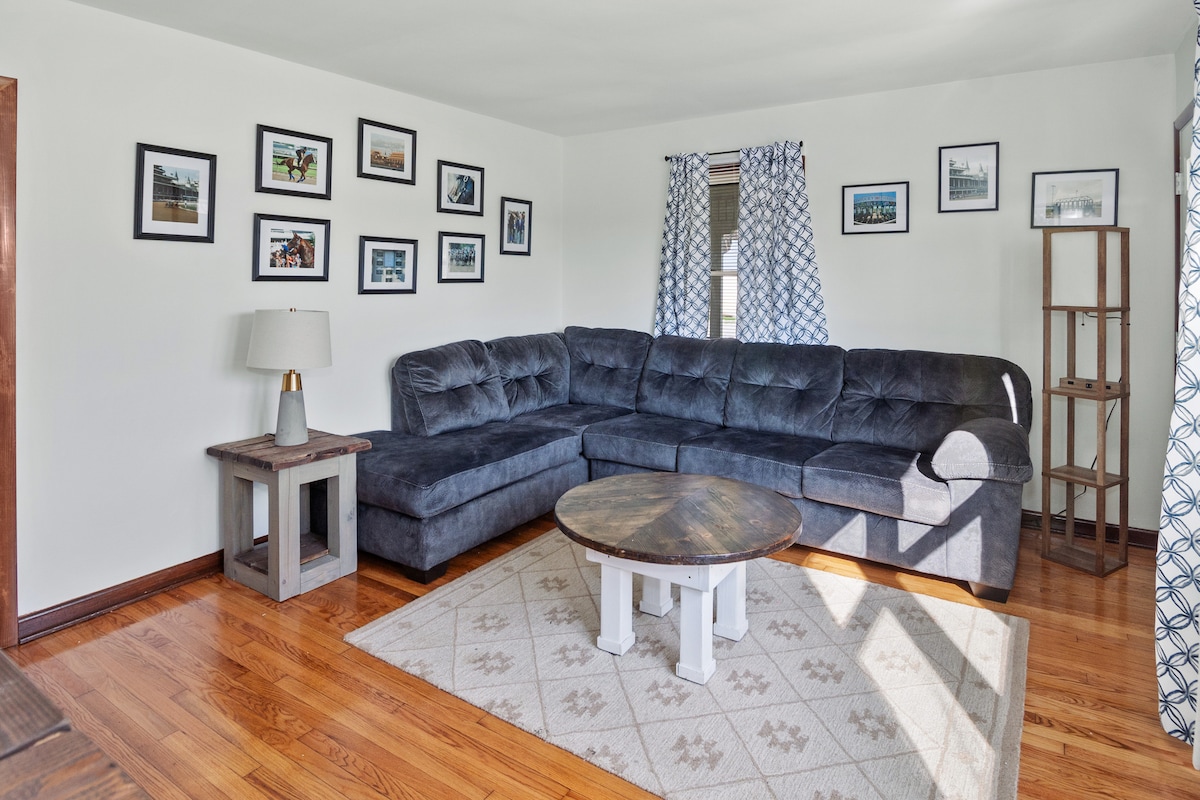 A spacious living room features a dark sectional couch arranged around a circular coffee table. Framed photographs adorn the walls, adding character. Natural light filters through patterned curtains, illuminating the hardwood floors and a neutral area rug underfoot.
