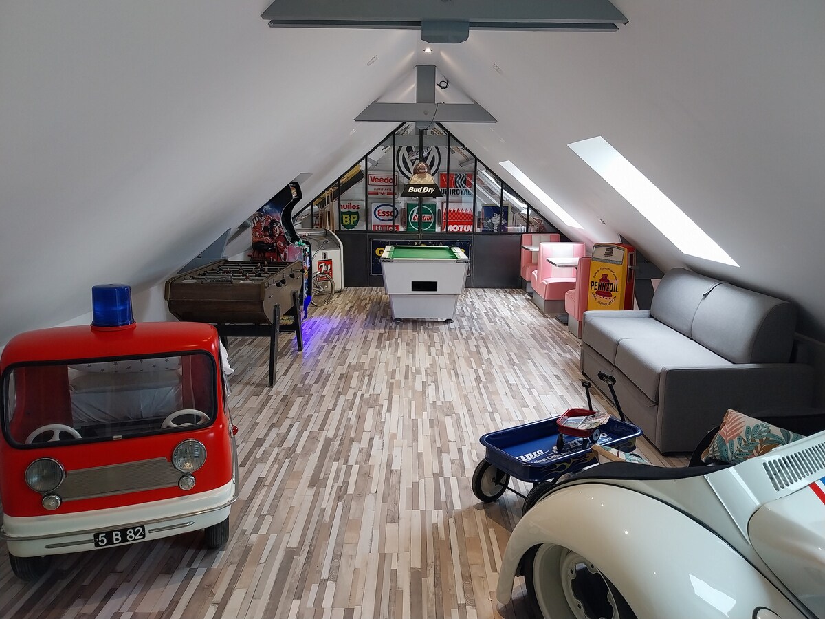 An upper-level game room is presented, featuring a billiards table, a foosball table, and a retro arcade machine. A grey sofa is positioned against one wall. Two toy cars, including a vintage-style model, are placed on the floor, surrounded by wooden flooring and skylights for natural light.