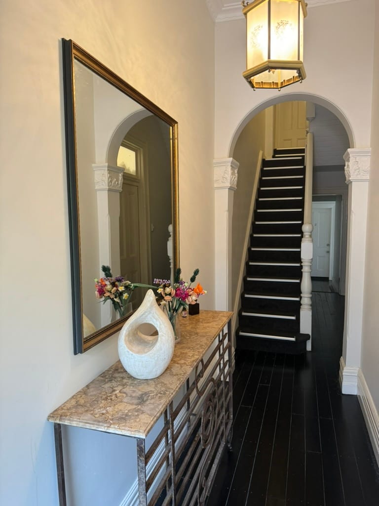 A stylish entryway is highlighted by a decorative console table with a marble top. A large mirror reflects the warm light from an elegant overhead fixture. An archway frames the staircase, where dark wood steps lead to the upper level, creating a welcoming atmosphere.