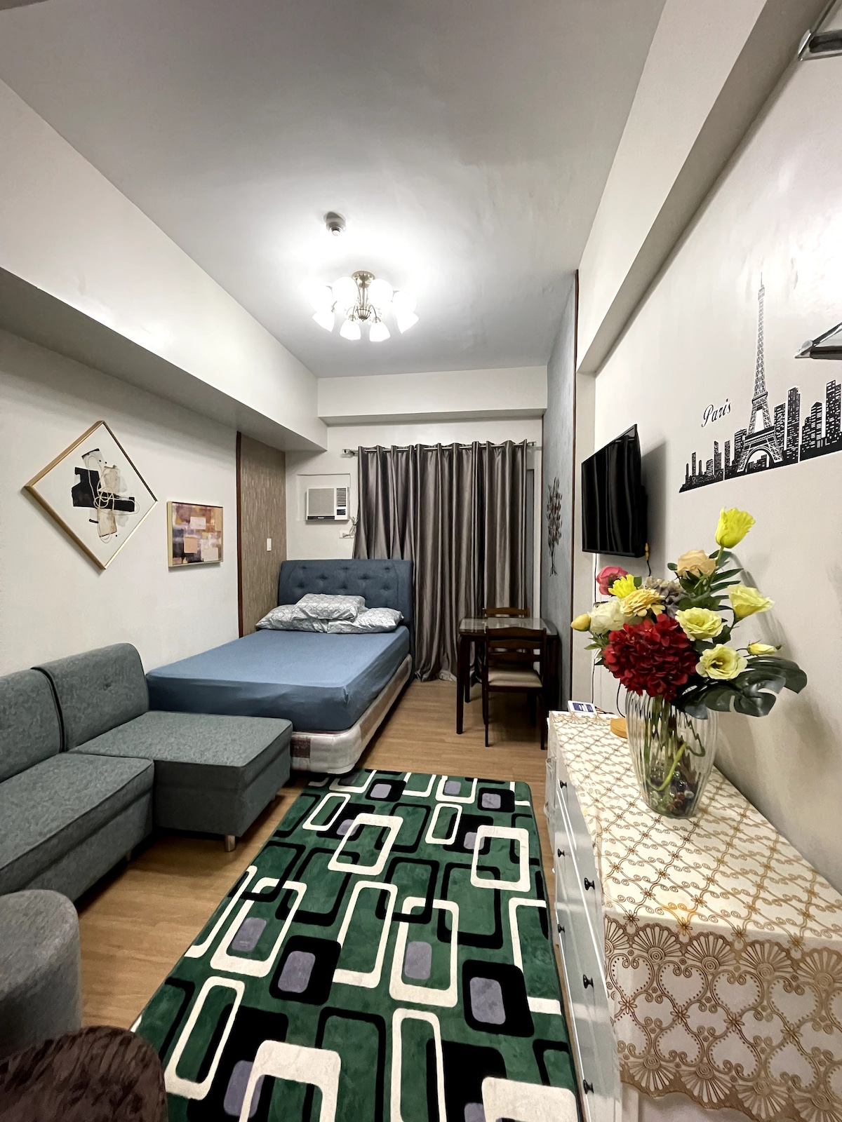 A cozy room is seen with a spacious bed and a sitting area furnished with a gray couch. Decorative artwork adorns the walls, while a vibrant rug adds color to the wooden floor. A television is mounted near the window, which is covered by elegant curtains.