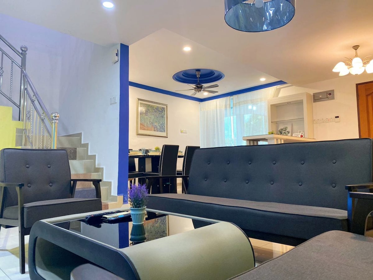 A cozy living room is shown with modern, dark sofas arranged around a glass coffee table. A small potted plant sits on the table. Bright lighting enhances the airy atmosphere, complemented by a ceiling fan and decorative wall art. A dining area is visible in the background.