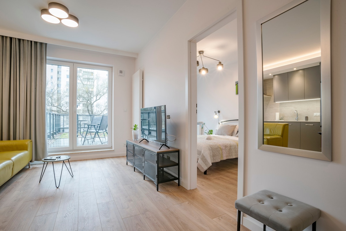 A welcoming living space is highlighted by a yellow sofa and a sleek coffee table. Glass doors lead to a balcony, allowing natural light to fill the room. A mirror reflects the cozy area, which connects to a bedroom and modern kitchen.