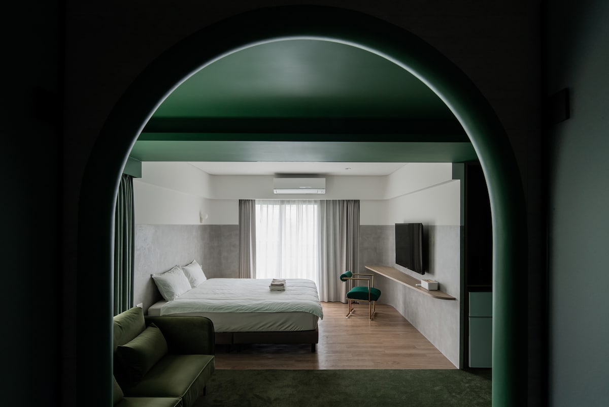 A spacious room is framed by an arched entrance, revealing a bright area with a queen bed and a cozy green sofa. Natural light filters through sheer curtains, illuminating the minimalist décor and accent features, including a TV mounted on a light-colored wall.