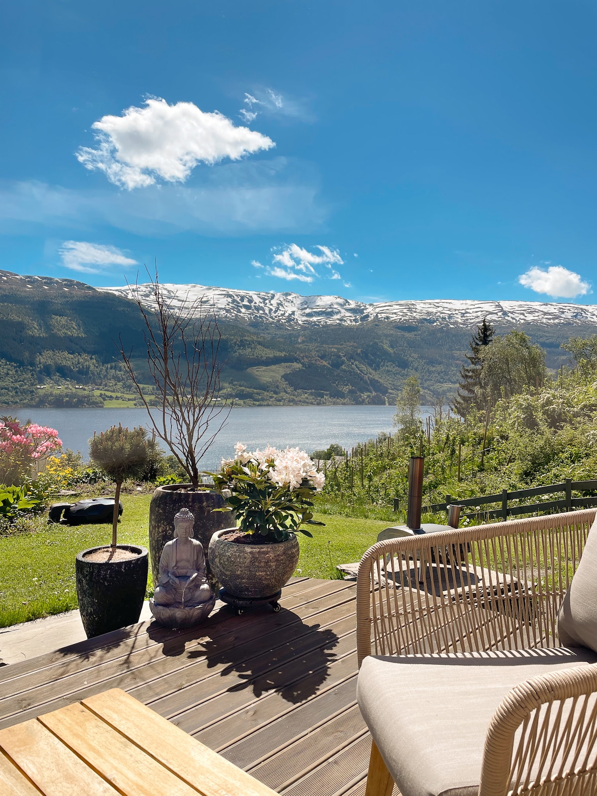A serene outdoor space is presented, featuring a wooden deck with comfortable seating. Potted plants and a stone statue offer a calming presence. In the background, mountains with snow-capped peaks rise above a sparkling lake under a clear blue sky.