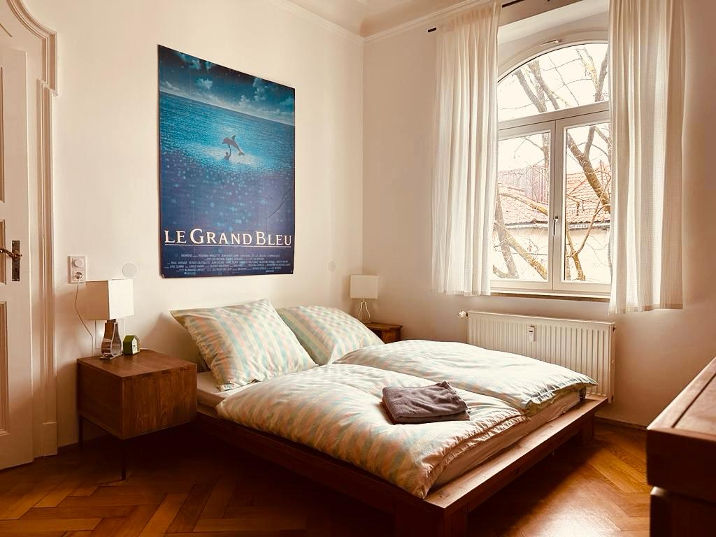 A bright bedroom features a spacious bed with patterned bedding. A poster is displayed above the bed, and light spills in through a large window adorned with sheer curtains. Two bedside tables are positioned on either side, each with a lamp.