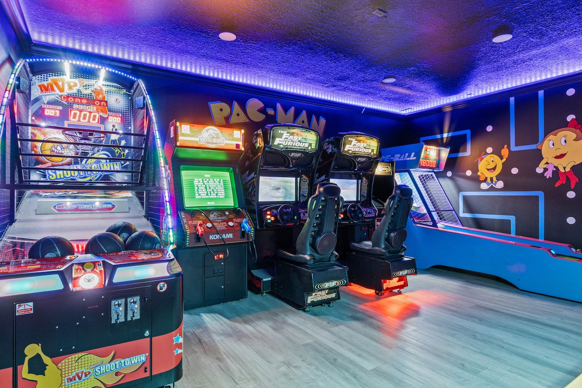 An inviting game room is showcased with a variety of arcade machines, including arcade classics like Pac-Man. Bright neon lights outline the room, enhancing a sense of excitement. A mix of shooting and racing arcade games, along with an air hockey table, creates a vibrant atmosphere for entertainment.
