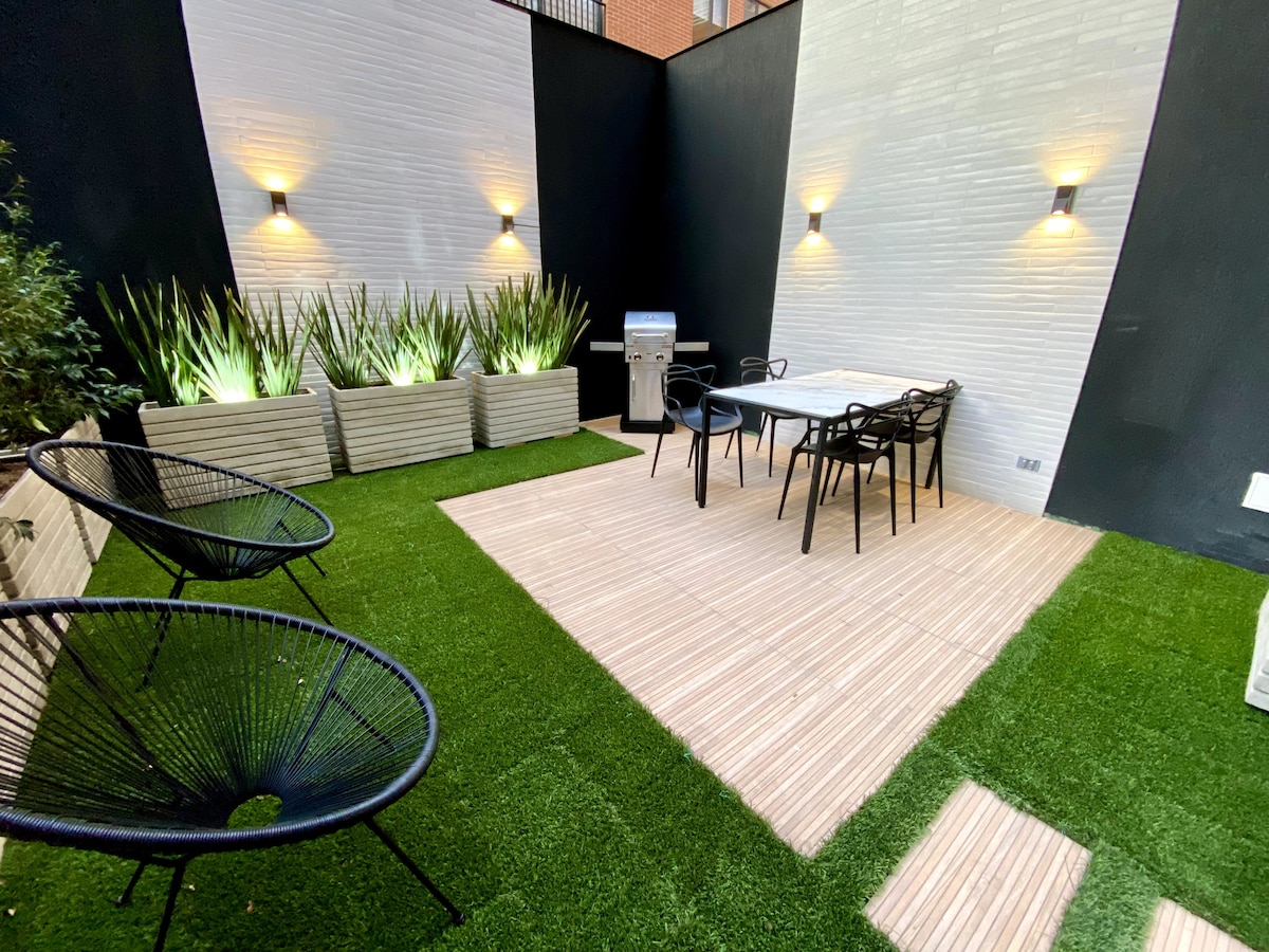 A private patio features a well-maintained grassy area surrounded by modern planters with tall greenery. A dining table with black chairs is positioned alongside a barbecue grill, with soft lighting from wall sconces illuminating the space for evening gatherings.