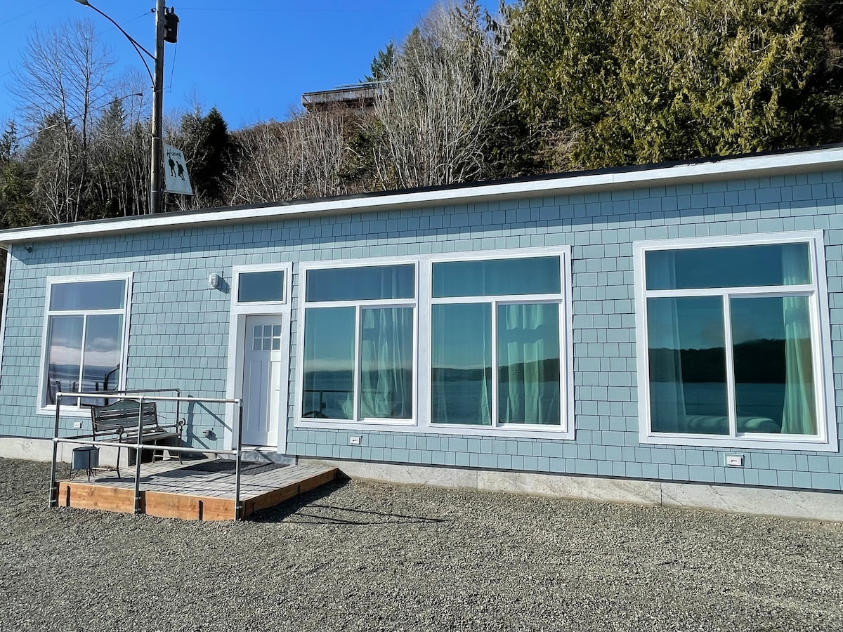 A single-story cottage features large windows that offer expansive views of the waterfront. Natural light fills the space, highlighting the tasteful exterior in light blue. A wooden deck with seating is visible, providing a welcoming entrance to this tranquil getaway.