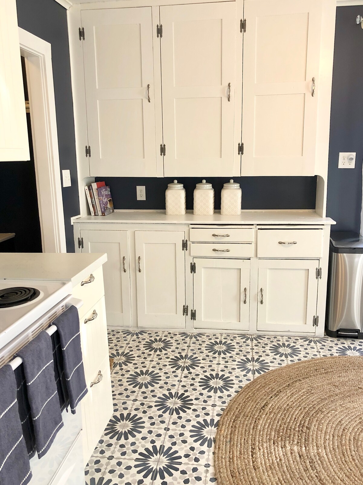 A clean and functional kitchen space is highlighted by white cabinetry and a patterned tile floor. The countertop is arranged with three clear containers, and kitchen towels are neatly hung on the stove. A round area rug adds texture to the room.