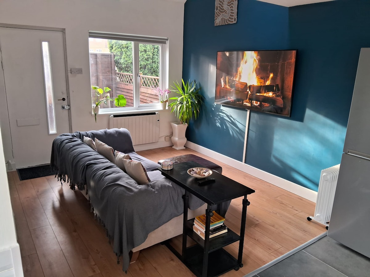 A cozy seating area features a comfortable sofa draped with a soft throw, positioned near a wall-mounted television displaying a crackling fireplace. A small black table holds decorative items and books. Natural light enters through a large window, illuminating the warm tones of the space.