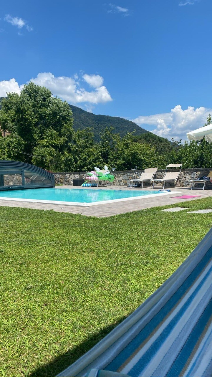 An open green lawn frames a salt-water pool, reflecting the blue sky above. Lounge chairs are positioned around the pool, and a colorful inflatable floats on the water's surface. A scenic backdrop of mountains adds to the tranquil setting.