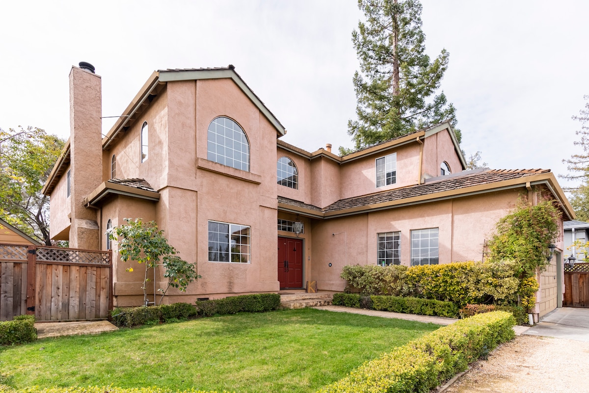 A two-story house is presented with a warm-toned exterior, featuring large arched windows that allow natural light to fill the space. Lush landscaping surrounds the front yard, complete with neatly trimmed hedges and a pathway leading to the double front doors.