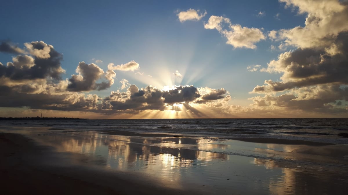 The beach showcases a stunning sunset, with golden rays emerging from behind clouds. The sun's light reflects gently on the wet sand, creating a serene atmosphere. Soft waves can be seen lapping at the shore, while the sky transitions from blue to warm hues.