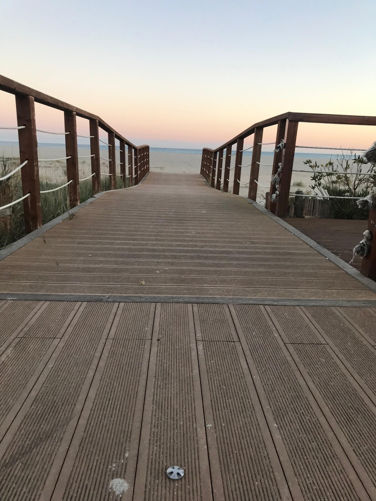 A wooden walkway leads directly to the sandy beach, with wooden railings on either side. The beach stretches toward the horizon, where the sky exhibits soft pastel colors at dusk, transitioning from warm tones to cooler shades as the sun sets.