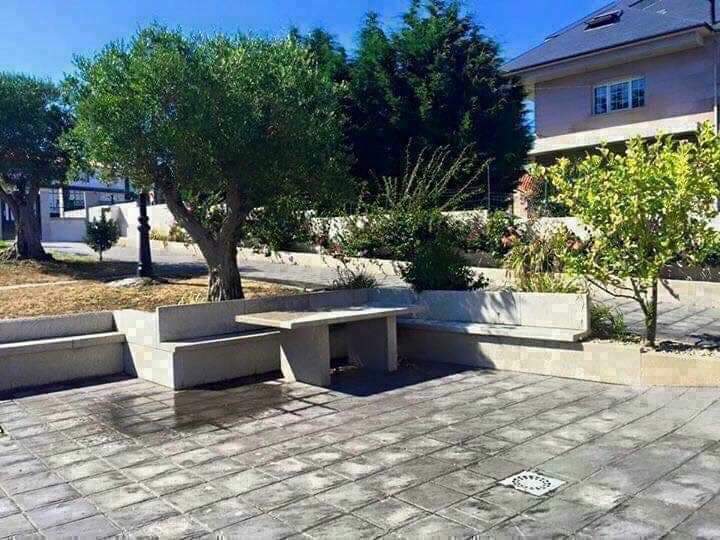 A spacious outdoor area features a stone bench arrangement and a small table set on a tiled surface. Lush greenery and trees provide natural shade, while the surroundings are well-maintained, creating an inviting space for relaxation.