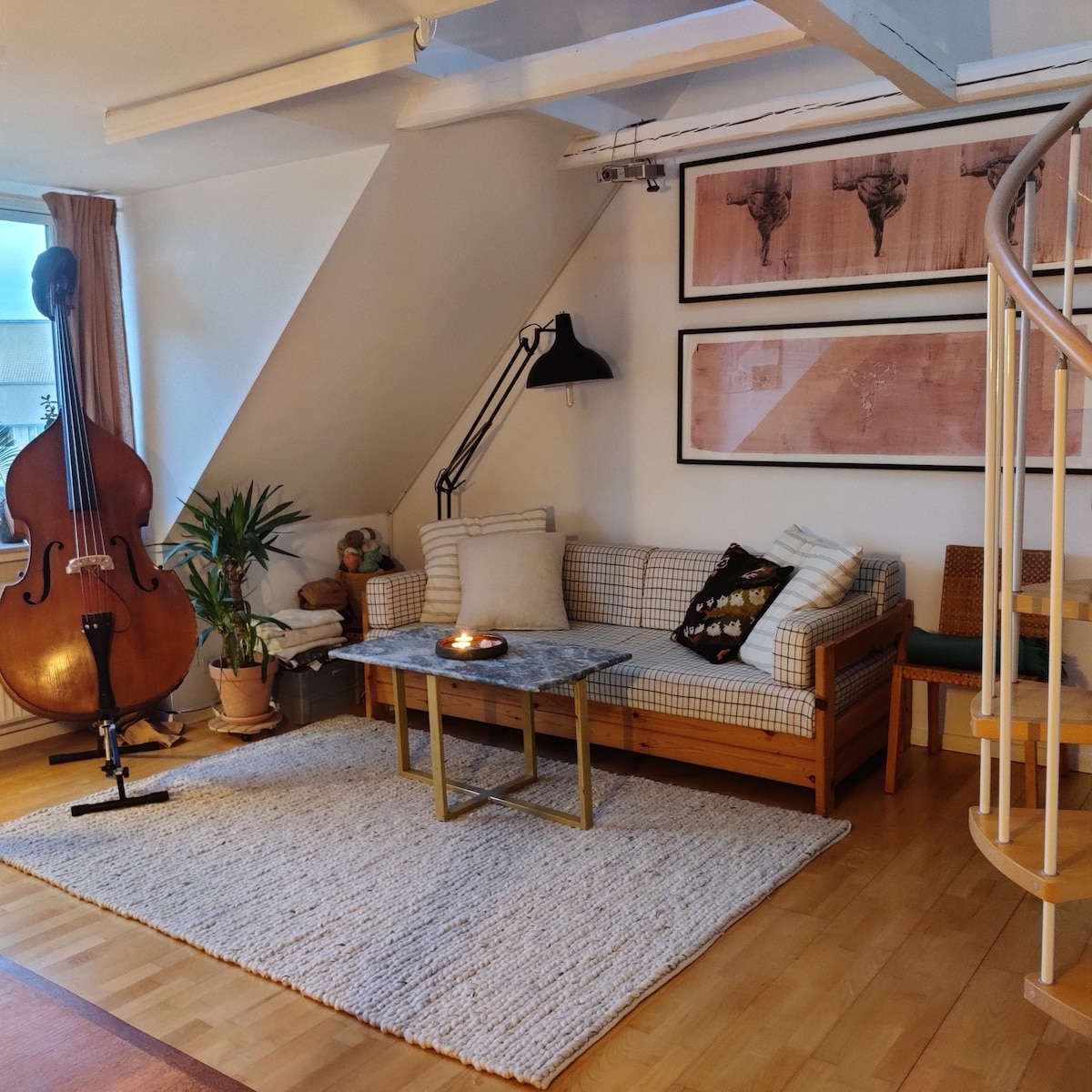 A cozy living area is showcased, featuring a pull-out sofa adorned with patterned cushions and a marble coffee table. A large musical instrument stands near the wall, complemented by plants and framed artwork, with soft light filtering through a window.