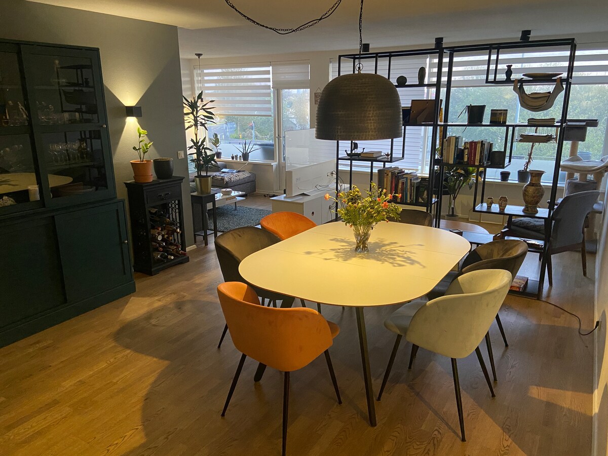 A modern dining area features a large, round white table surrounded by six stylish chairs in various colors. Natural light fills the space through large windows, while a decorative pendant light hangs above. A bookshelf and plants add visual interest to the backdrop.