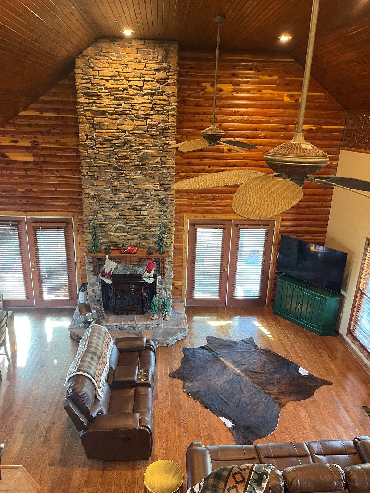 The great room features a stone fireplace accented with holiday stockings. Two leather sofas are arranged around a decorative cowhide rug. Large windows provide natural light and views of the outdoors, while a nearby TV sits on a low green cabinet.