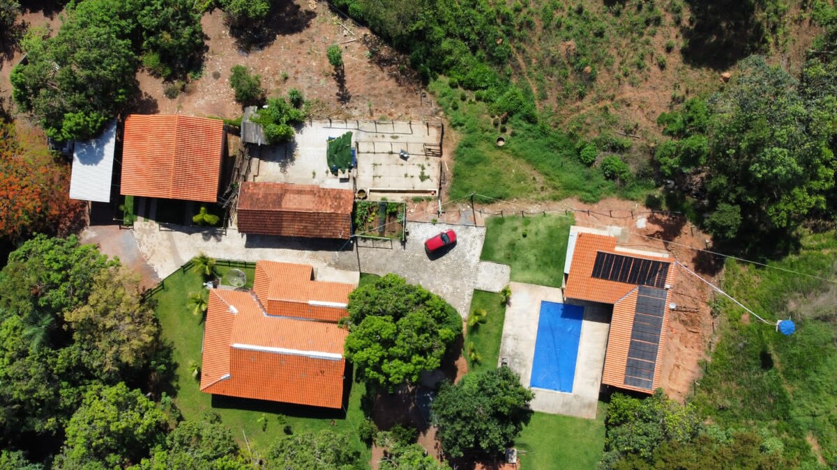 An aerial view of a serene property highlights multiple buildings with terracotta roofs surrounded by lush greenery. A blue swimming pool is visible alongside a red car parked on a stone path, while gardens and outdoor areas are seen nearby.