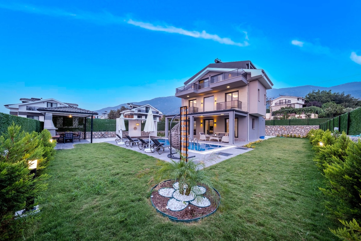 A modern three-story villa is showcased against a clear blue sky. The spacious yard features a well-maintained lawn, outlined by green hedges. Sun loungers are positioned near the pool area, with a patio set visible on the side, creating an inviting outdoor space.