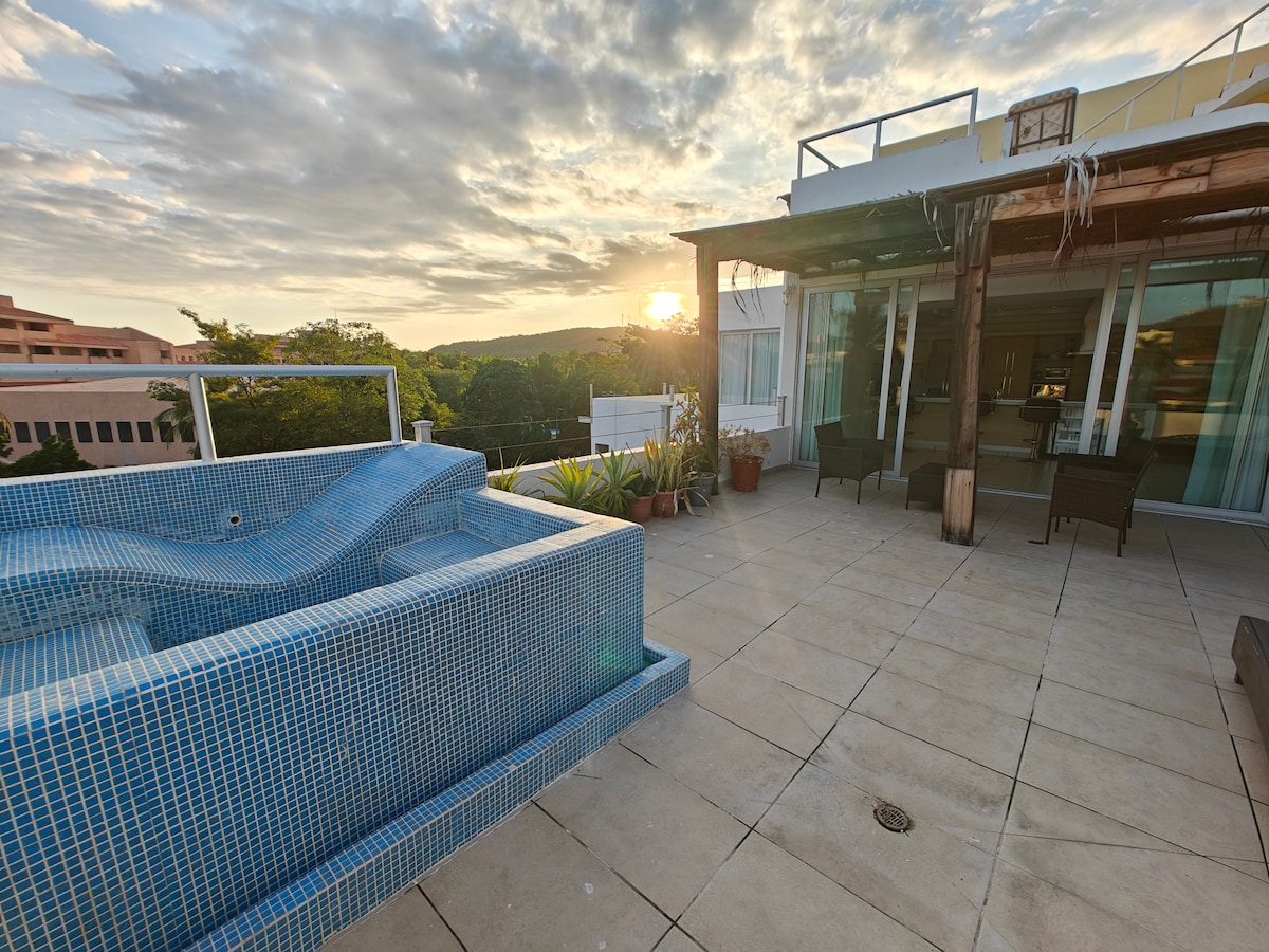 A spacious terrace features a tiled hot tub, surrounded by potted plants. Comfortable seating areas are arranged for relaxation, with a view of the setting sun illuminating the sky. Natural light highlights the nearby glass doors leading into the interior space.