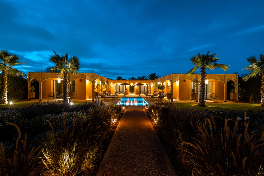 The villa is illuminated at night, showcasing a long pathway leading to a serene pool. Flanking the pool, palm trees and softly lit buildings create a harmonious ambiance. The surrounding garden features lush plants, enhancing the tranquil outdoor setting.
