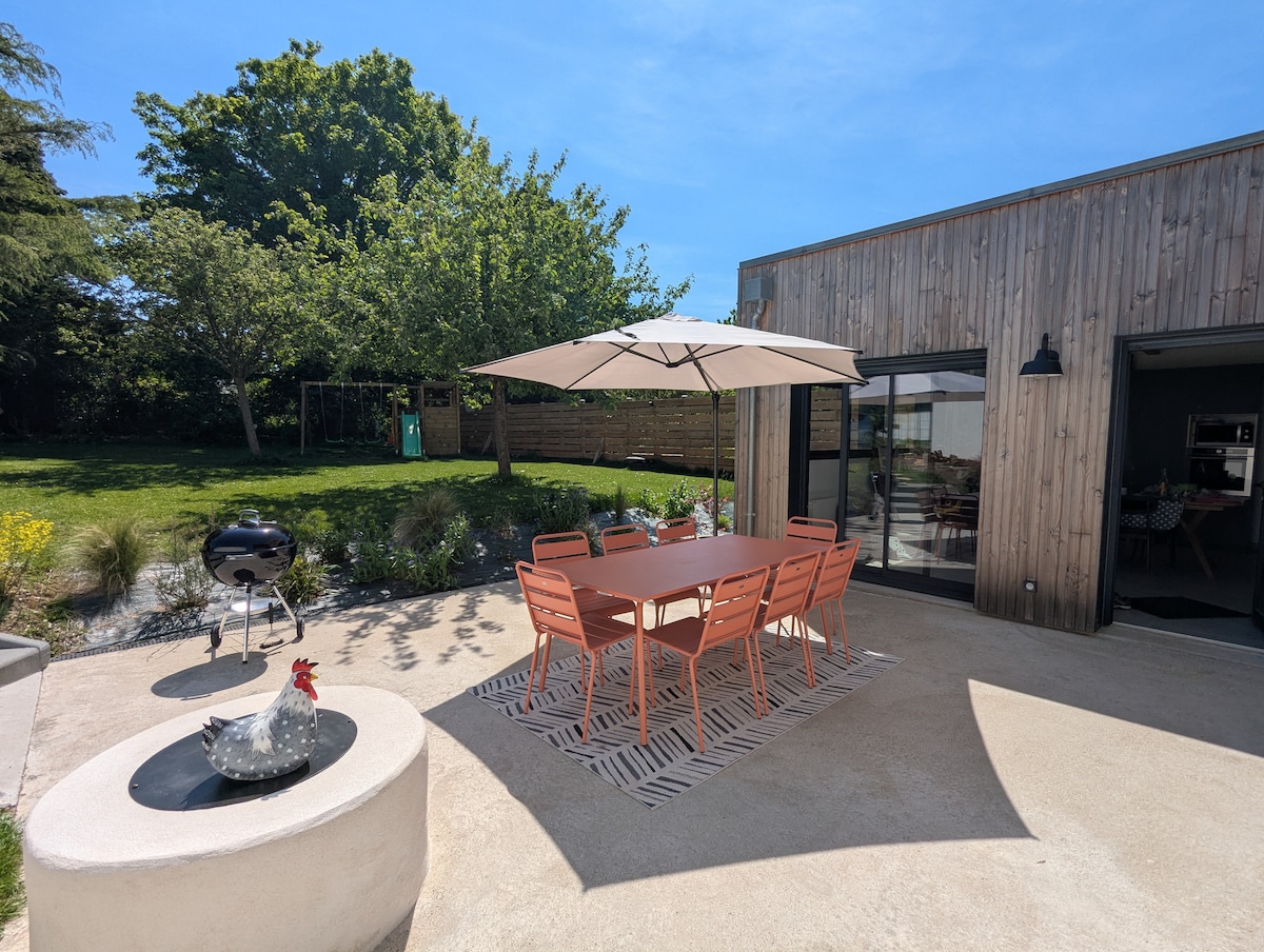 An outdoor dining area is presented with a large table surrounded by six coral-colored chairs. A large umbrella provides shade over the table, while a barbecue grill and decorative elements are visible nearby. Lush greenery frames the serene setting.
