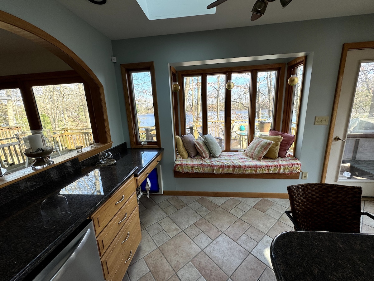 A bright kitchen space features a skylight and a window seat with colorful cushions, providing views of the surrounding wooded area and the lake. The granite countertops, wooden cabinetry, and appliances create a functional atmosphere, seamlessly connected to the outdoor scenery.
