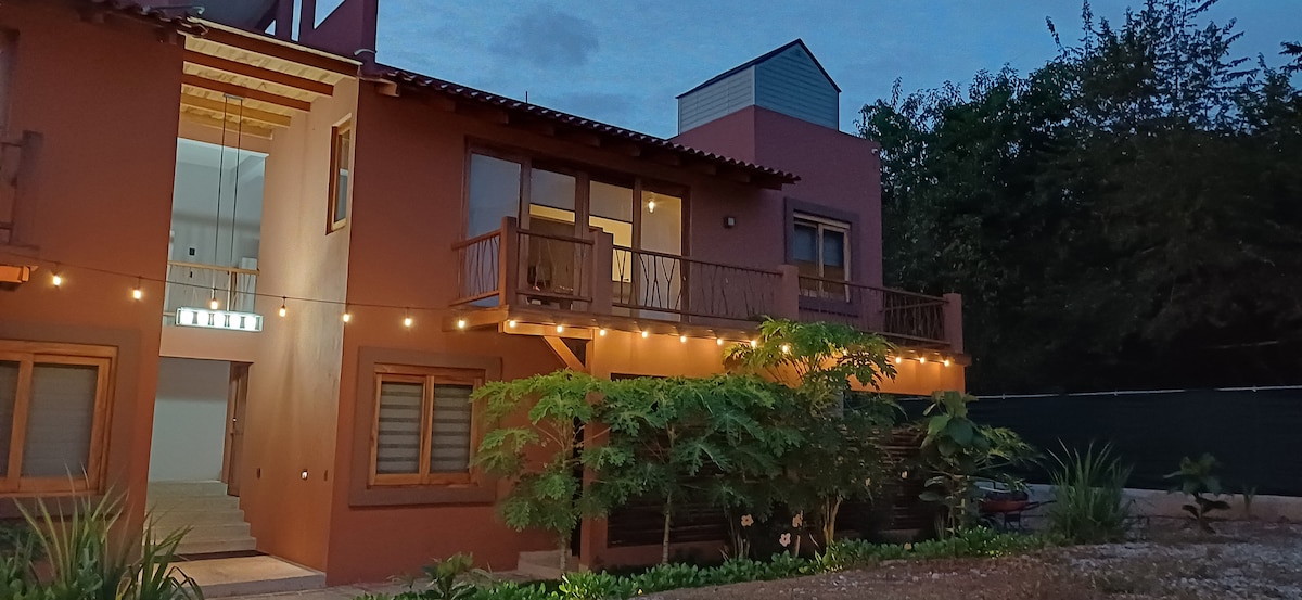 A two-story building is set against a dusky sky, featuring wooden balconies adorned with soft lighting. Green plants frame the entrance, while large windows create an open, inviting atmosphere. The warm exterior colors offer a sense of comfort in this tranquil environment.