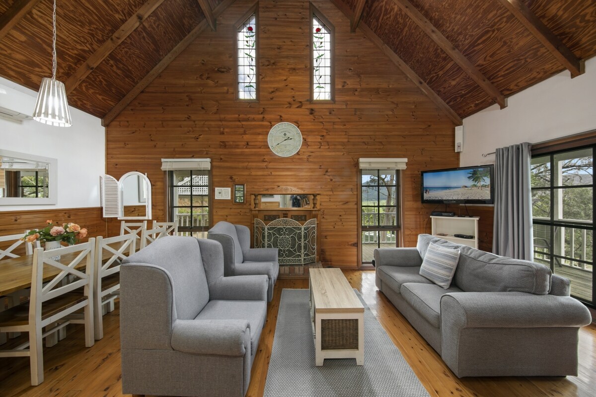 An open-plan lounge and dining area features high ceilings with exposed wooden beams. Two comfortable grey sofas are arranged around a central coffee table. A dining table with white chairs is visible, alongside a television and a fireplace, all under large windows allowing natural light.