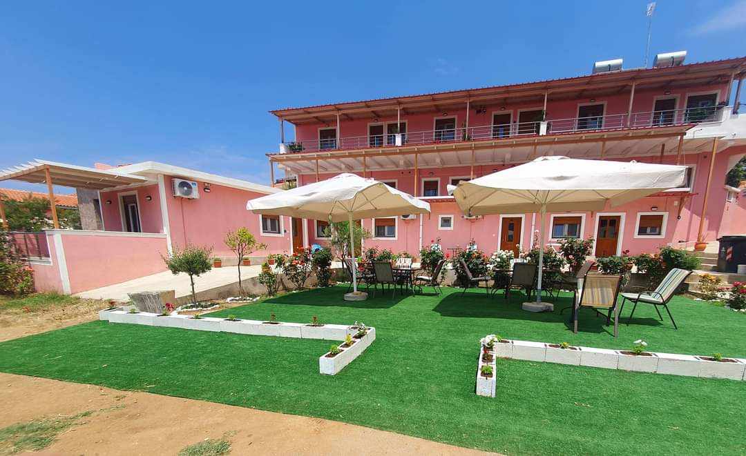 A vibrant outdoor area is presented, featuring white umbrellas shading several tables and chairs on lush green turf. Flower beds with blooming plants surround the seating area. The pink building offers a welcoming backdrop, complete with several windows and balconies.