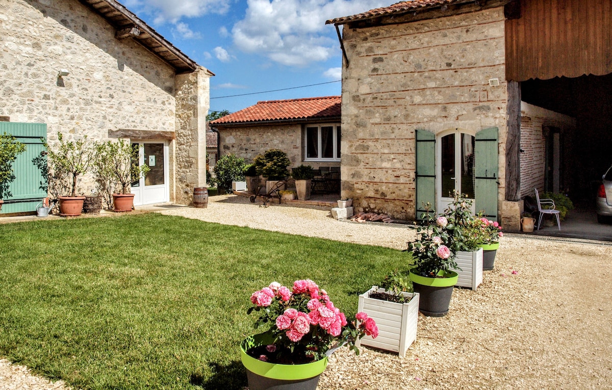 The image captures a serene courtyard featuring three stone buildings with vibrant green shutters. Potted flowers in shades of pink are arranged along a gravel path, leading towards grassy areas. A blue sky with fluffy clouds enhances the peaceful rural setting.