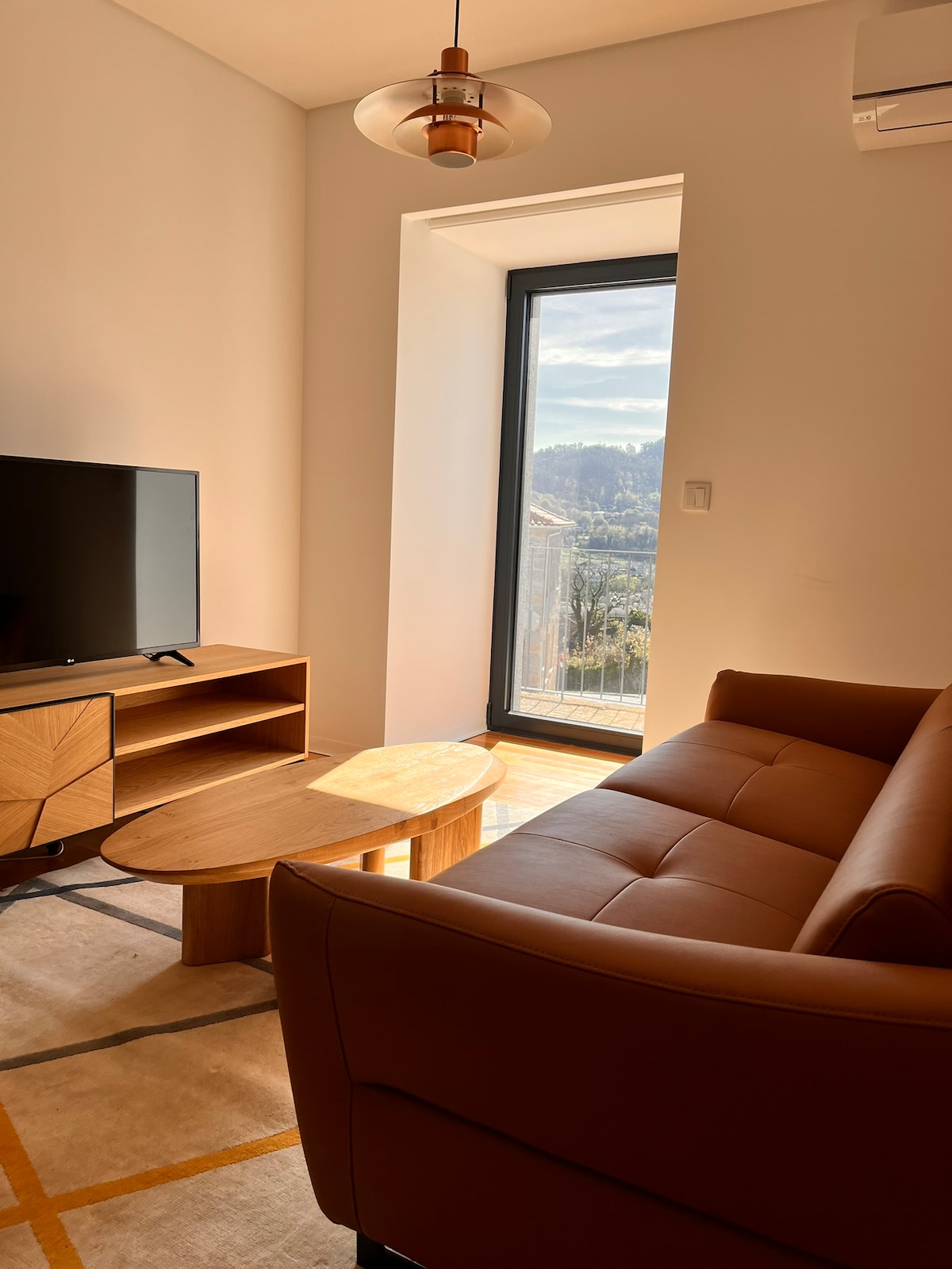 A stylish living area is featured with a modern brown sofa and a wooden coffee table. A television sits atop a matching wooden console. Natural light floods the space through a large window, offering a view of the scenic outdoors.