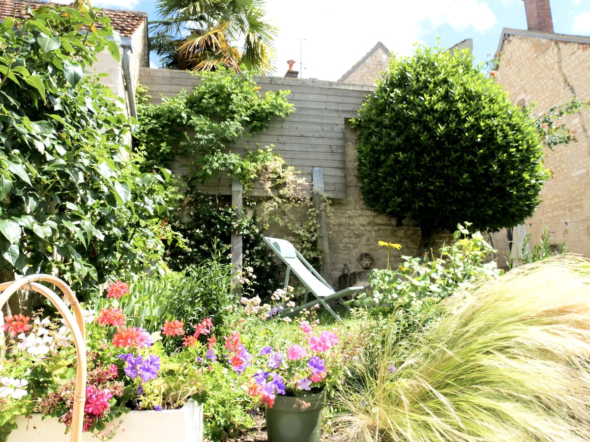 A vibrant garden is showcased, featuring a variety of colorful flowers in bloom. A green lounge chair is positioned among the lush vegetation and decorative shrubs, while a tall bush provides shade. The scene is complemented by a wooden wall in the background.