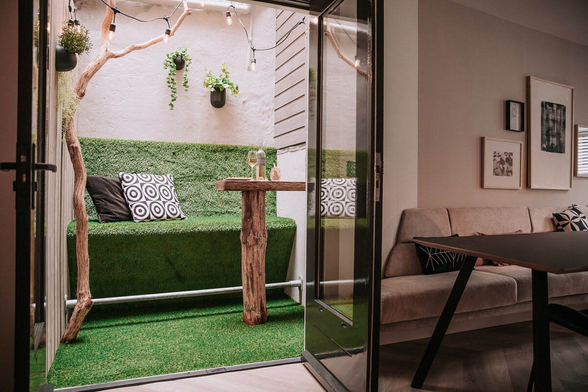 An inviting outdoor space is captured, featuring a cozy seating area with a green grass-like surface. A small table made from natural wood complements the setting. Soft lighting strings hang overhead, enhancing the relaxed atmosphere. A nearby wall displays hanging plants for added greenery.