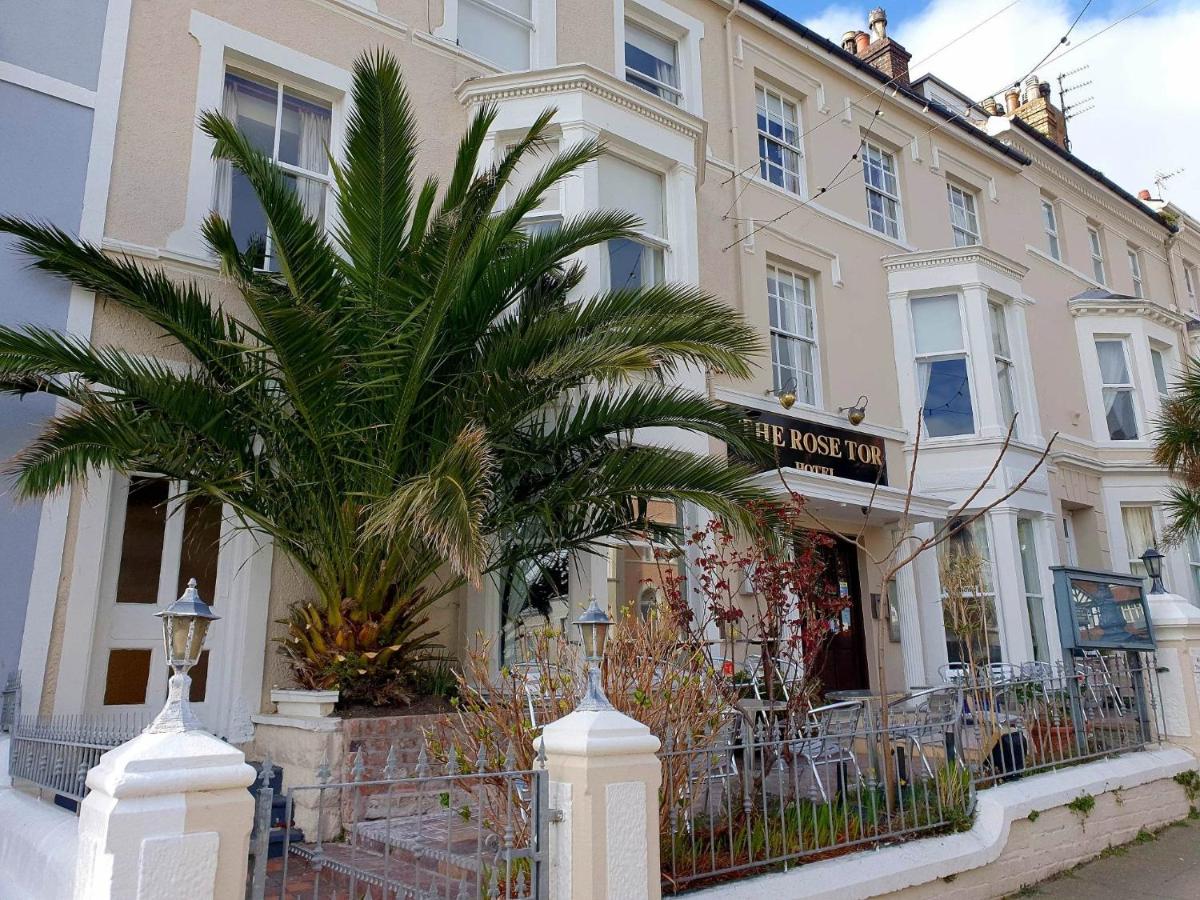The exterior of the Rose Tor Hotel features a light-colored façade with multiple large windows. Lush greenery, including a palm tree, is positioned prominently in front. A decorative wrought-iron fence lines the property, contributing to the charming appearance of the building.