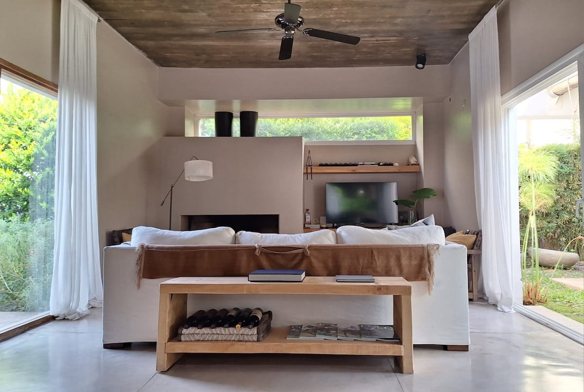 A spacious living area features a cozy white couch, complemented by a wooden coffee table with a selection of books. Large windows allow natural light to fill the space, viewed with greenery outside. A sleek television sits against a muted wall, contributing to the calming environment.