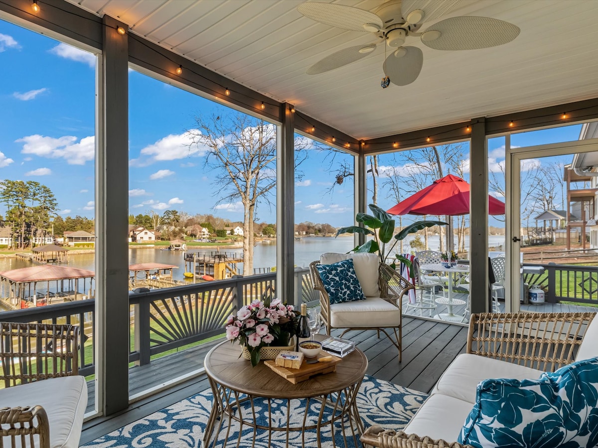 A screened porch offers comfortable seating with a round table at its center. Decorative plants and a floral arrangement add natural elements. Large windows frame scenic views of the lake and surrounding area, while a red umbrella provides shade for outdoor enjoyment.