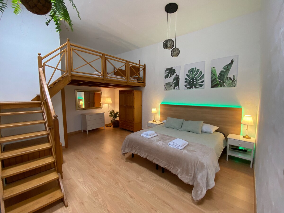 A spacious bedroom features a large bed with soft bedding. A wooden loft overlooks the area. Light fixtures cast a warm glow, while tropical leaf artwork adorns the wall above the bed. A staircase provides access to the loft, enhancing the room's open layout.